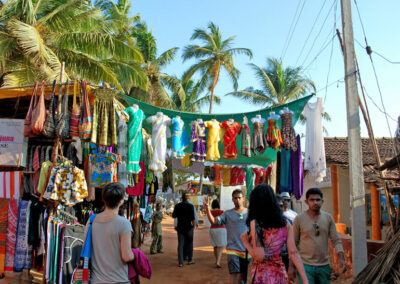 Eine Marktgasse unter Palmen. Zu beiden Seiten stehen Markstände mit bunter Ware wie Taschen und Kleidung. Quer über der Gasse hängen bunte indische Kleider. Mehrere Personen sind zu sehen.