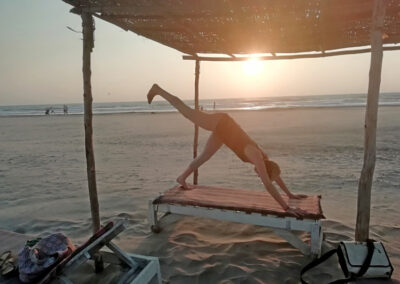 Abendliche Stimmung am fast menschenleeren Strand. Eine Person übt die Yoga-Pose "Herabschauender Hund" auf einer Holzliege, die sich unter einer Überdachung aus Schilfmatten befindet.