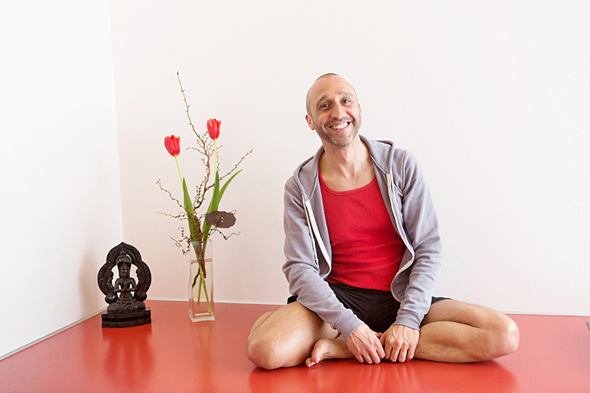 Matthias Kärcher sitzt auf dem roten Linoleumfußboden seines Yogastudios. Neben ihm auf dem Boden stehen eine kleine Patanjali-Statue und eine Vase mit zwei roten Tulpen.