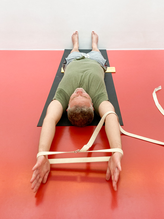 Ein Yogaschüler liegt mit dem Rücken auf der Yogamatte. Beine und Arme sind gestreckt, die Fußsohlen berühren die Wand. Die Arme liegen in Verlängerung des Körpers am Boden, die Handgelenke drücken in die Schlaufe eine Gurtes.