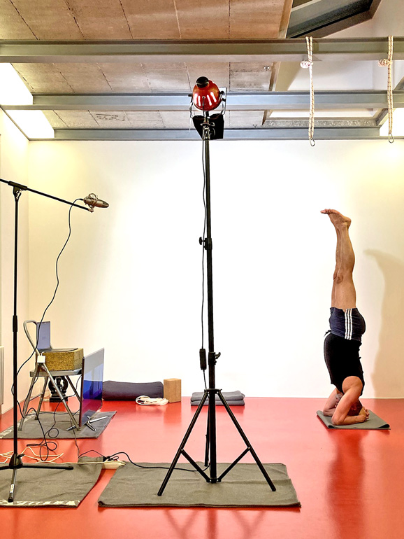 Szene aus dem Online-Unterricht im Yoga-Studio. Ein Scheinwerfer auf einem Stativ beleuchtet Matthias, der einen Kopfstand macht. Ihm gegenüber steht ein großer Flachbildschirm auf dem Boden, links im Bild befindet sich ein Mikrofon auf einem zweiten Stativ.