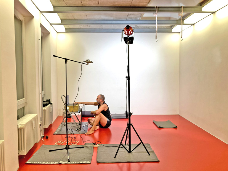 Blick in ein Yogastudio. Im Vordergrund stehen zwei Stative, am linken Stativ ist ein Mikrofon befestigt, am rechten ein Scheinwerfer. Im Hintergrund sitzt Matthias auf dem Boden, die eine Hand auf der Tastatur eines Laptops, das auf einem Stuhl platziert ist.