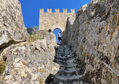 Blick auf die ausgetretenen Stufen einer steinernen Treppe, die wie in den Felsen gehöhlt steil nach oben zu einem zinnenbekrönten Tor führt. Im Durchgang steht eine Person und schaut nach unten.