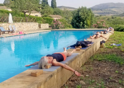 Sechs Personen ruhen in der Schlussentspannung nach dem Yoga in Rückenlage auf dem breiten Rand eines Pooles.