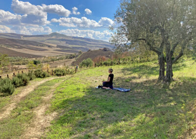 Seitenansicht einer einzelnen Frau, die auf einem Yogakissen im Schatten eines Baumes sitzt und in die Hügellandschaft vor ihr schaut.