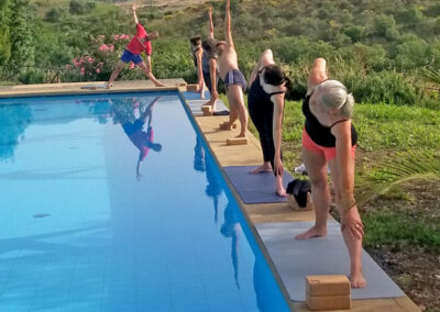 Sechs Personen üben Yoga auf dem mattenbreiten Seitenrand einen blauen Swimmingpools. Im Hintergund befindet sich eine karge Hügellandschaft mit niedrigen Bäumen.