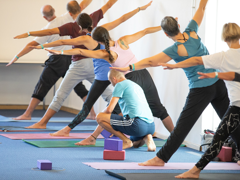 Sieben Yogaschüler stehen hintereinander aufgereiht mit gegrätschten Beinen und neigen den Oberkörper mit seitlich ausgestreckten Armen nach links. Der Yogalehrer kniet neben einer Schülerin und korrigiert deren Beinausrichtung mit einem Yogaklotz.