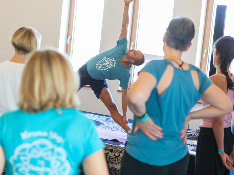 Rückansicht von vier Workshop-Teilnehmerinnen, die dem Lehrer Matthias dabei zuschauen, wie er eine Yoga-Stellung ausführt.