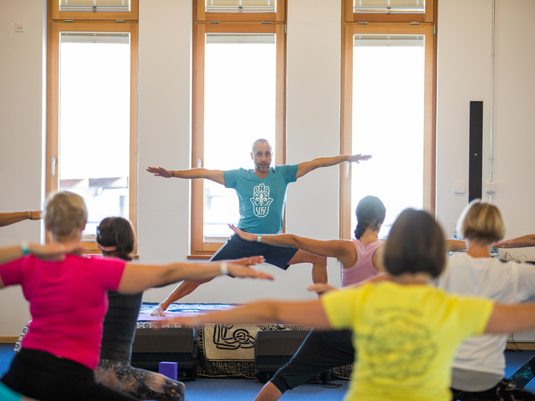 Rückansicht von Yogaschülern, die gemeinsam mit dem Lehrer eine Yoga-Pose ausführen. Der Lehrer steht dabei erhöht auf einem Podest, hinter ihm sind drei schmale Fenster.