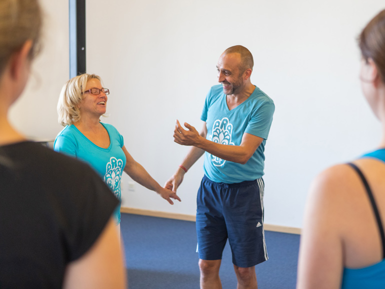 Matthias und eine Workshop-Teilnehmerin in einer humorvollen Situation. Sie stehen einander gegenüber und lachen, Matthias gestikuliert und hält hält den nach unten getreckten Arm der Frau. Zwei Frauen im Bildvordergrund beobachten die Szene.