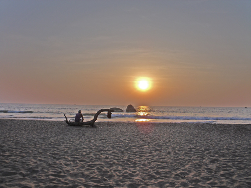 Person sitzt auf Treibholz am Sandstrand und blickt auf Sonnenuntergang über dem Meer mit Felsen im Wasser.