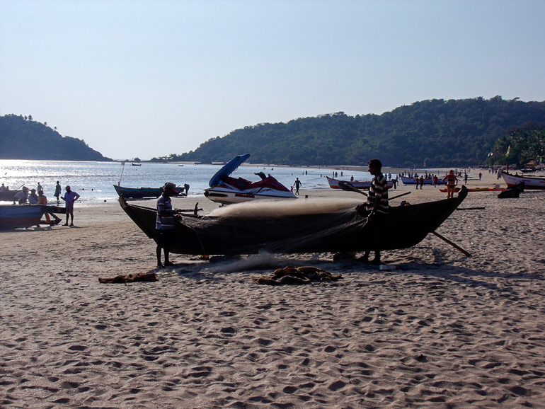 Zwei Fischer mit Boot und Netz am Strand von Palolem in Goa, im Hintergrund das Meer und Besucher.