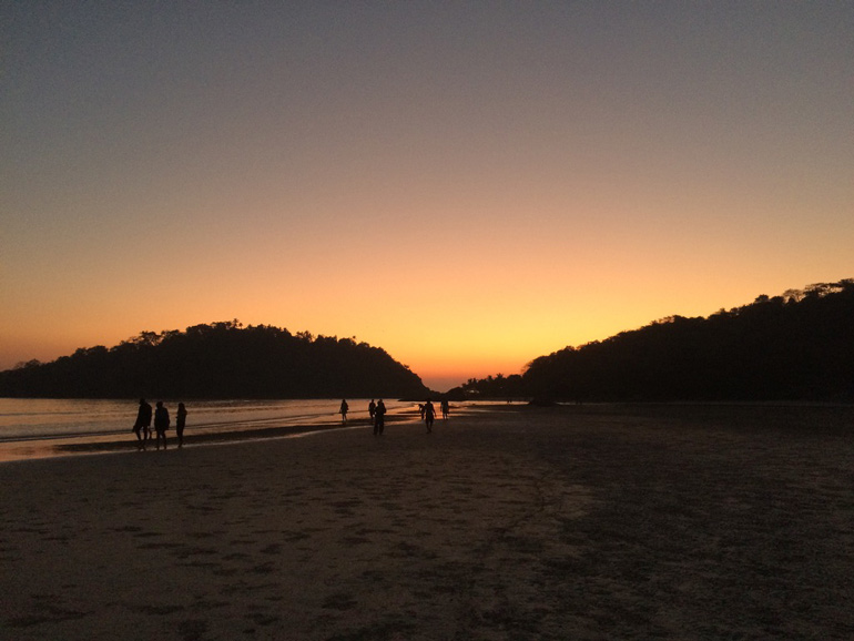 Menschen spazieren am Palolem Beach bei Sonnenuntergang, mit orange-violettem Himmel über dem Meer.