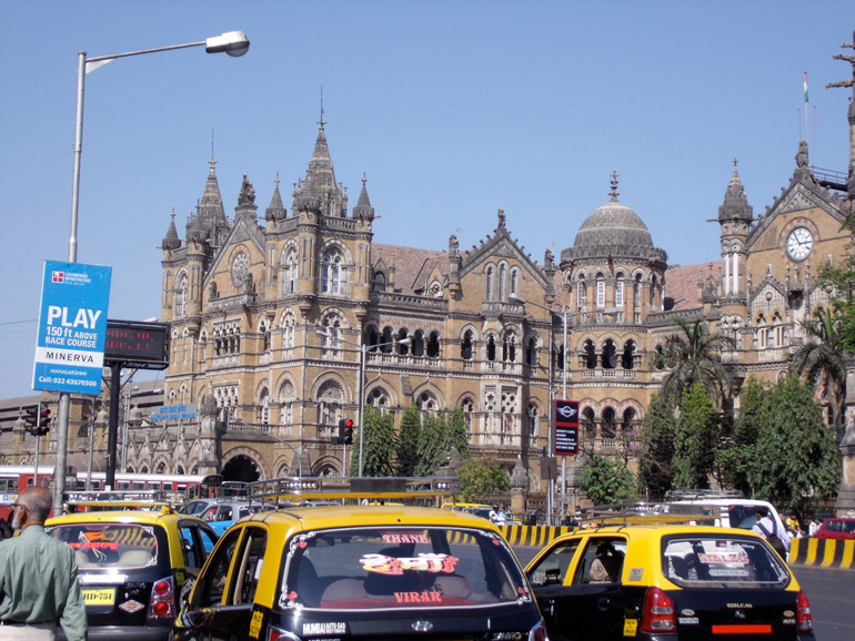 Historischer Bahnhof Chhatrapati Shivaji Terminus in Mumbai mit gelben Taxis im Vordergrund.
