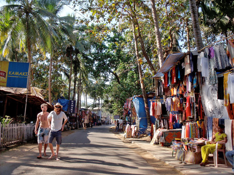 Bunte Marktstraße in Goa mit Palmen, Kleidungständen und zwei Touristen beim Flanieren.
