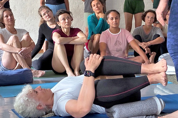 Im Vordergrund eine Yoga-Schülerin liegend, mit angewinkelten Beinen; dahiner sitzend andere Workshop-Teilnehmer.