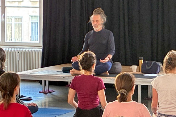 Yoga-Lehrer im Mediationssitz auf einem Podest, vor im sitzende Yoga-Übende.